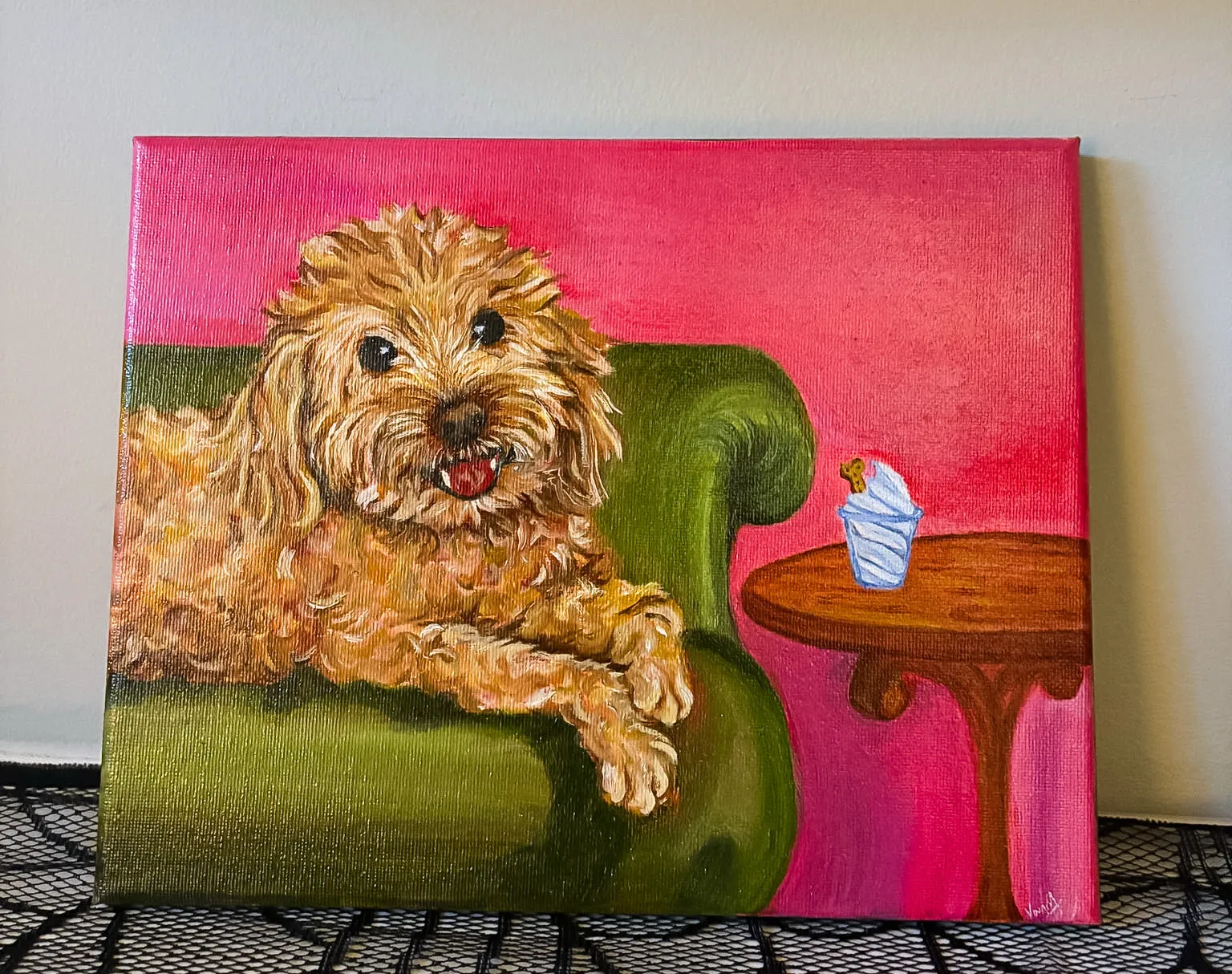 The finished canvas propped against a pale wall on a black lace cloth — a curly golden dog lounging on a green sofa beside a small wooden table against a hot-pink background.